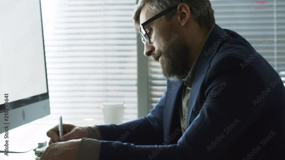 Poster handheld shot of middle-aged man in glasses writing in notebook when having phone conversation at of