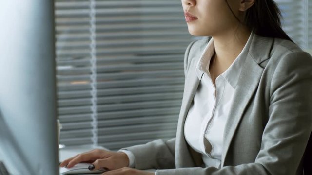 Handheld shot of concentrated Asian businesswoman working with desktop computer when sitting at desk in office