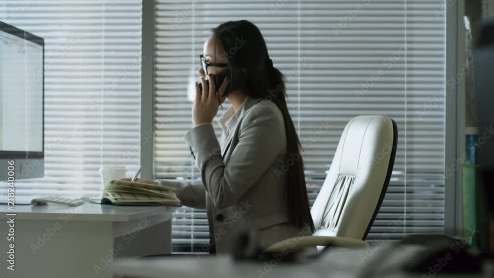 Poster side view of young asian woman in glasses and formal clothes talking on the phone when working with 