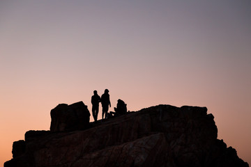 Silhouette of teens handing out atop a rock at sunset.