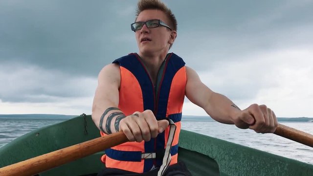 A young man in a boat rowing wooden oars on the water. Life jacket on the body of a man.