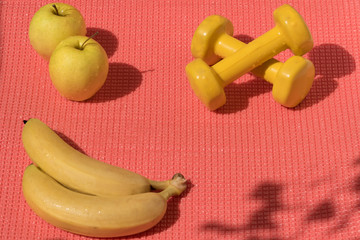 Fototapeta premium Healthy lifestyle in modern colors: Two yellow dumbbells, apples, banans on the pink exercise matt. Shadows cast by sunlight on the surface.