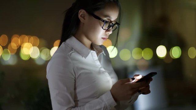 Side view of young Asian woman wearing white shirt and glasses using mobile phone when standing by window late at night
