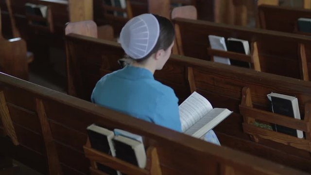 A Mennonite woman sits in an empty church pew and flips through a hymnal.