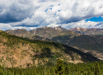 Sunny, evergreen foreground with overlapping Colorado Rocky Mountains beyond.  Dark clouds, but a hint of blue sky.  Good for background, text.