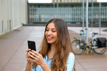 Smiling young woman is using an app in her smartphone device to send a text message while standing in courtyard of office blocks