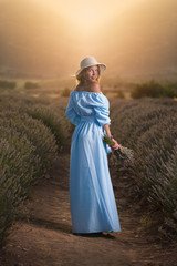 Brunette young woman in a blue dress with a bouquet of lavender in a lavender field during sunset