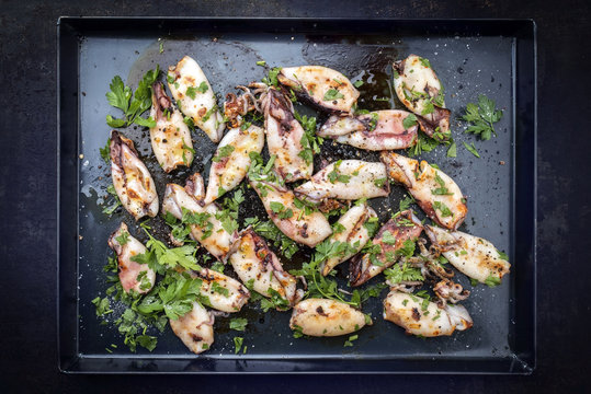 Traditional barbecue Greek calamari with herb and spices as top view on a black metal sheet