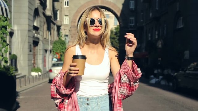 Young blonde wowam walking at the steet and drinking coffee. Steadycam shot.