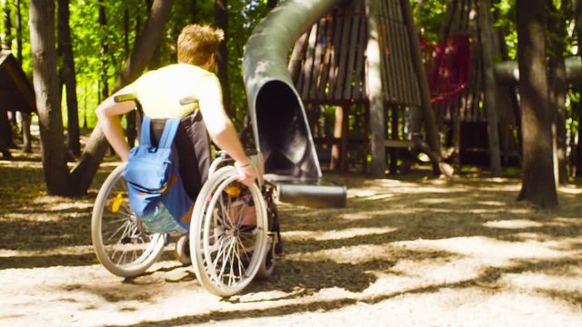Dolly shot. Happy young disable man in a wheelchair in the park. He is riding near rope playground.