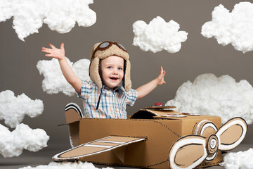 the boy plays in an airplane made of cardboard box and dreams of becoming a pilot, clouds from cotton wool on a gray background, retro style