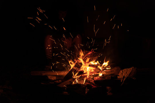 A Wood Fire Blazes In A Fire Pit As Family Members And Friends Gather Around At Night To Roast Marshmallows