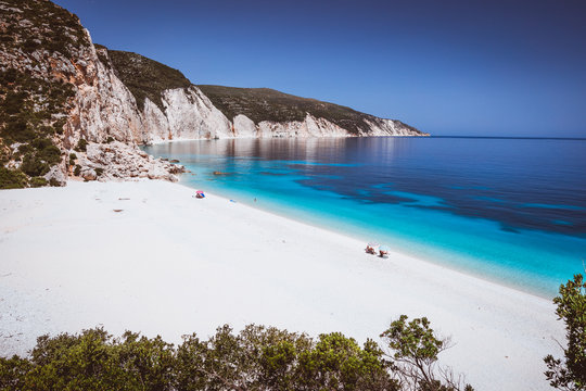 Fteri beach, Kefalonia, Greece. Lonely tourists protected from sun umbrella chill relax near clear blue emerald turquoise sea water lagoon. Framed by tree foliage