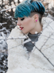 A nice girl with blue hair wrapped herself in a white fur coat in a snow-covered park. Rubbing against the background of a bush