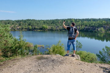 Fototapeta na wymiar Senior white man with backpack stands on the rock and making selfie at the lake background.