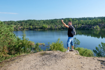 Fototapeta na wymiar Attractive senior white man with backpack stands on the rock and making selfie at the lake background.