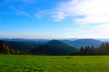 Hügelige Landschaft mit einem blauen Himmel und im Vordergrund eine grüne Wiese