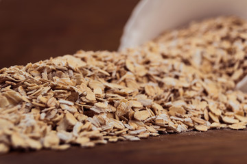 Oat flakes spilled out of a white porcelain bowl on the table, close-up, wooden background.