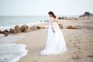 Fototapeta na wymiar A beautiful bride in a white wedding dress is walking along the beach. Waves near her dress. Beautiful sunset atmosphere, summer evening. Wedding concept.