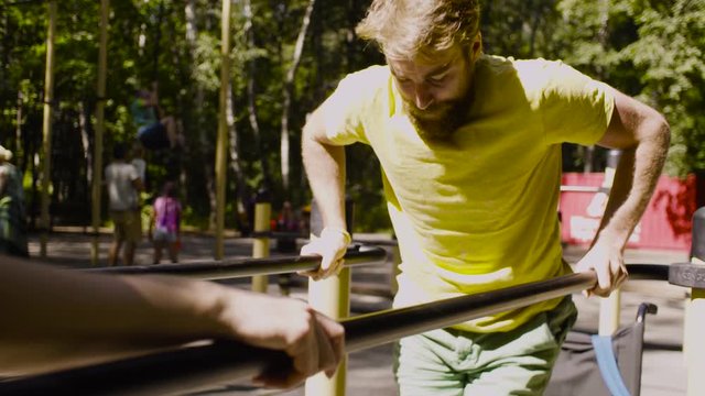 Young disable sportsman in the park with his wife. The man is doing exercises on the parallel bars, then the woman helping him.