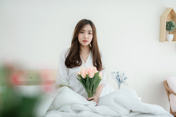 Obraz na płótnie Canvas Beautiful young woman holding a bouquet of tulips siting on white bed. Happy morning. Portrait of a smiling pretty young lady relaxing in white bed.