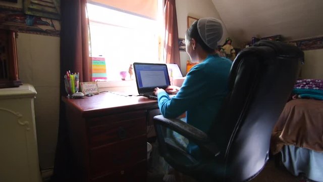 A young Mennonite woman smiling and talking as she works on a computer at her desk in slow motion.
