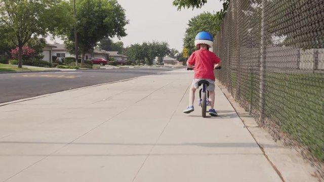 A young boy rides a blue balance bicycle along a sidewalk in a residential neighborhood.