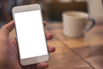 Mockup image of hand holding white mobile phone with blank desktop screen with coffee cup on wooden table in cafe
