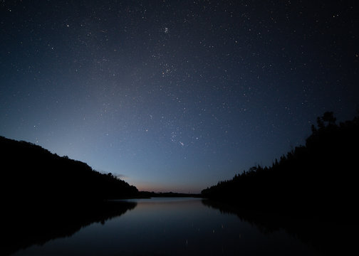 The fading stars in the early morning sky are reflected in a quiet lake and one small meteor from the Perseid meteor shower streaks across minutes before the daylight renders them invisible. 