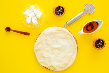 Raw rolled out pizza dough near cheese mozzarella, olive oil, spices, knife for pizza on yellow background top view mockup copy space