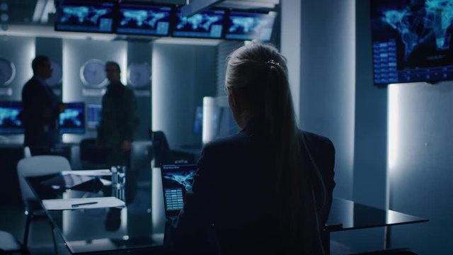 Female Special Agent Works on a Laptop in the Background Special Agent in Charge Talks To a Military Man in the Monitoring Room. In the Background Busy System Control Center with Monitors.