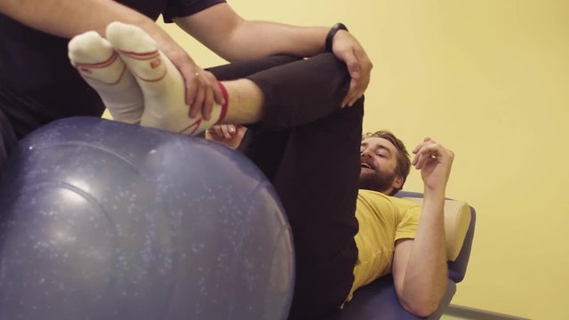 Young disabled man doing leg exercises at the rehabilitation center with a ball. Doctor physiotherapist helping him. Healthy gymnastics. Active people.