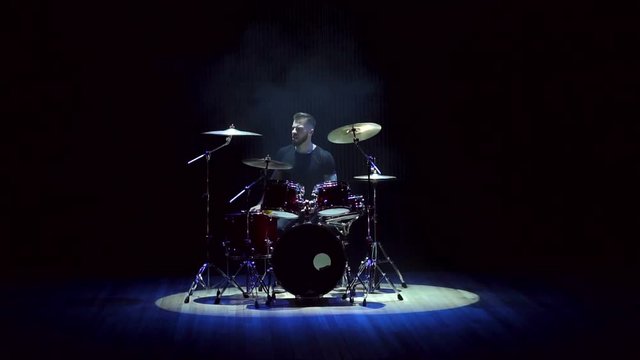 A young bearded man plays drums in the dark on stage during a concert, slow motion. The drummer is enjoying his performance during the concert. Man playing drums on black background with smoke.