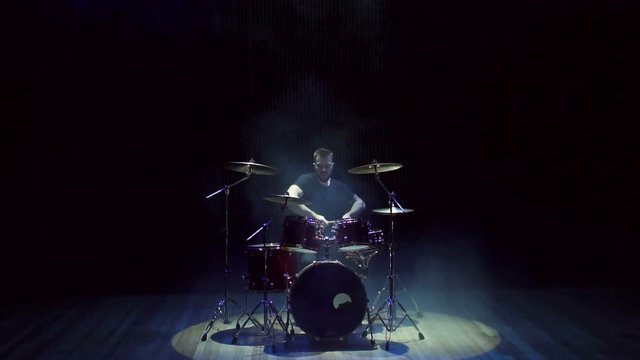 Active emotional guy plays drums on a bare stage in the dark, slow motion. Concert stage lighting with smoke. Male drummer artist musician playing the drums with drumsticks.