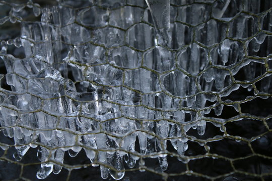 Close up macro of transparent frozen water icicled dripping hard in Winter extreme weather against a grid hexagon shape pattern garden pond cover in frost in icy morning outside in English countryside