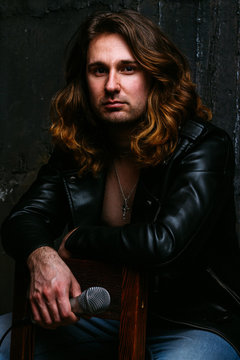 a portrait of a brutal, handsome man, a singer who sits on a chair on a dark background, in a black leather kurt on his naked body and holds a microphone in his hands. presenter, singer at the concert