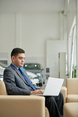Multi-ethnic owner of car dealership sitting on sofa and working on laptop