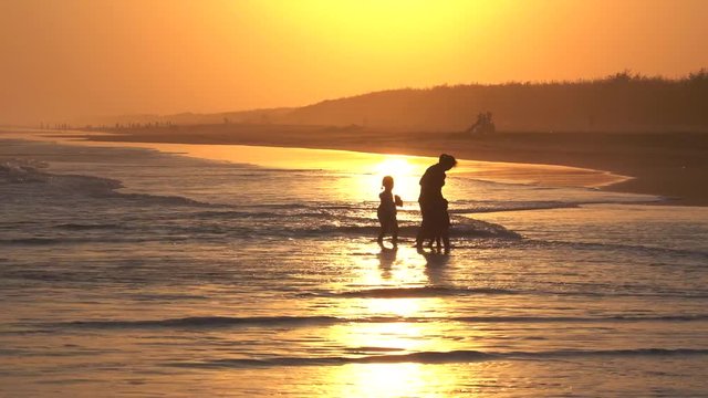 Yogyakarta, Indonesia - August 14, 2018: Silhouette of young mother and her children playing waves on the beach at sunset time. Shot in 4k resolution