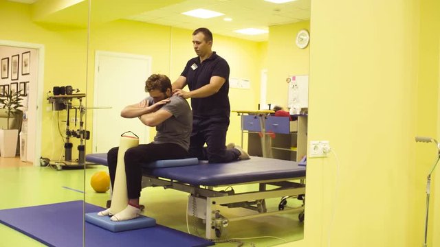 Doctor physiotherapist helping young disabled sportsman, doing exercises for a back at the rehabilitation center. Healthy gymnastics.