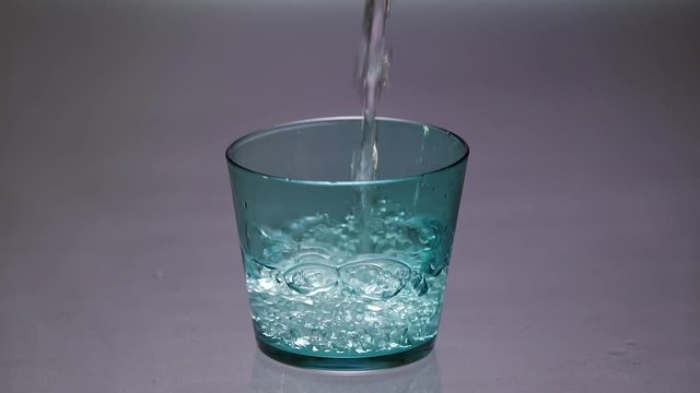 Pouring clear water into a blue glass until it overflows in slow motion.