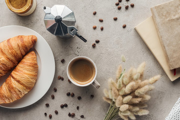 top view of cup of coffee with croissants and moka pot on concrete surface with lagurus ovatus bouquet and book