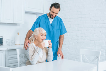 smiling male social worker looking at senior woman drinking tea in kitchen