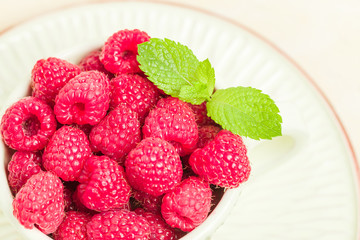 Ripe raspberries with green mint leaves in cup and saucer on pastel yellow background.