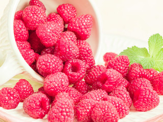Ripe raspberries with green mint leaves in cup and saucer on pastel yellow background.