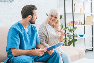 Obraz premium young male nurse holding clipboard and looking at smiling senior woman
