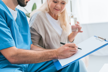 Fototapeta premium cropped shot of male nurse writing on clipboard and senior woman drinking water