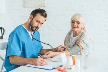 male nurse in stethoscope writing on clipboard while measuring blood pressure to senior woman