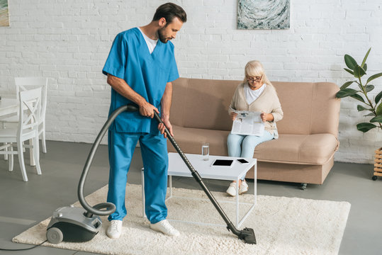 Young Social Worker Cleaning Carpet With Vacuum Cleaner While Senior Woman Reading Newspaper On Couch