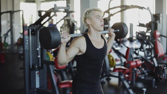 Sporty man holding barbell on the shoulder behind the neck while crouching and doing squats exercise in gym
