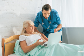 doctor with stethoscope looking at senior woman using laptop in hospital bed